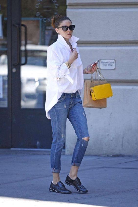 На девушке белая рубашка oversize, темно-синие джинсы с потертостями и черные полуботинки. Образ дополняют аксессуары, очки и желтая сумка.